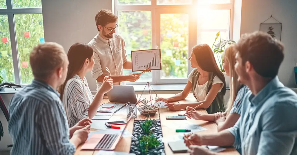 Corsi per lavorare Areajob- ragazzo che sta spiegando una sua strategia ai colleghi durante una riunione e sono tutti seduti al tavolo ad ascoltarlo e lui mostra un grafico su un dispositivo